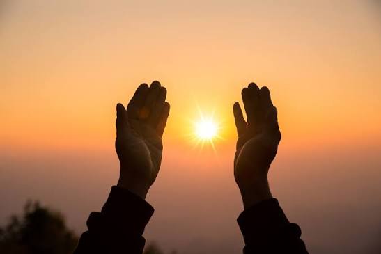 Mãos erguidas diante do sol em um céu alaranjado ao entardecer